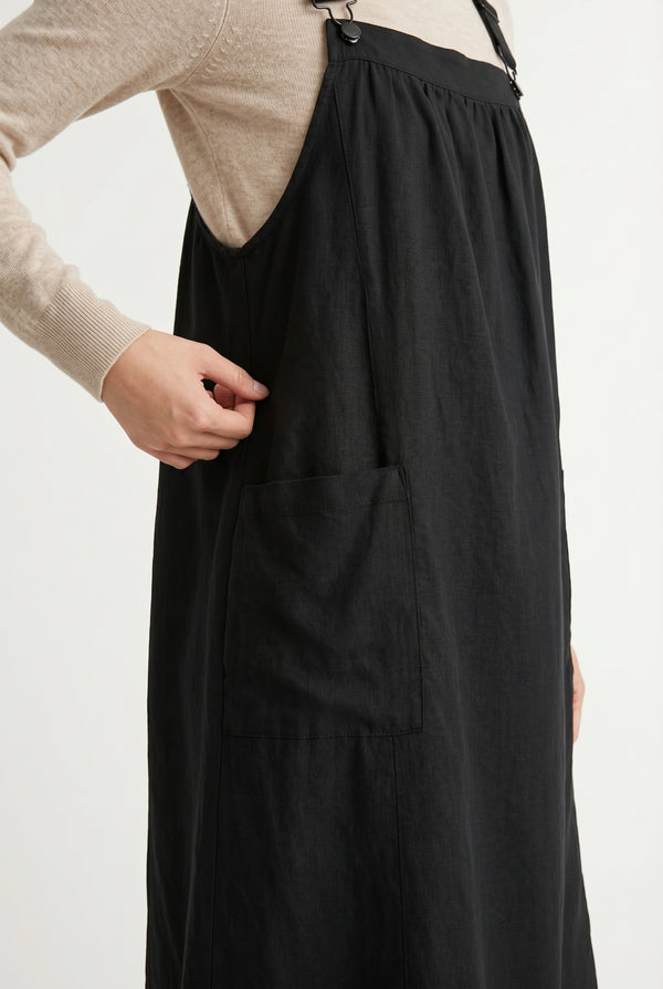 Woman wearing an onyx black midi-length pinafore dress layered over a white long-sleeve top