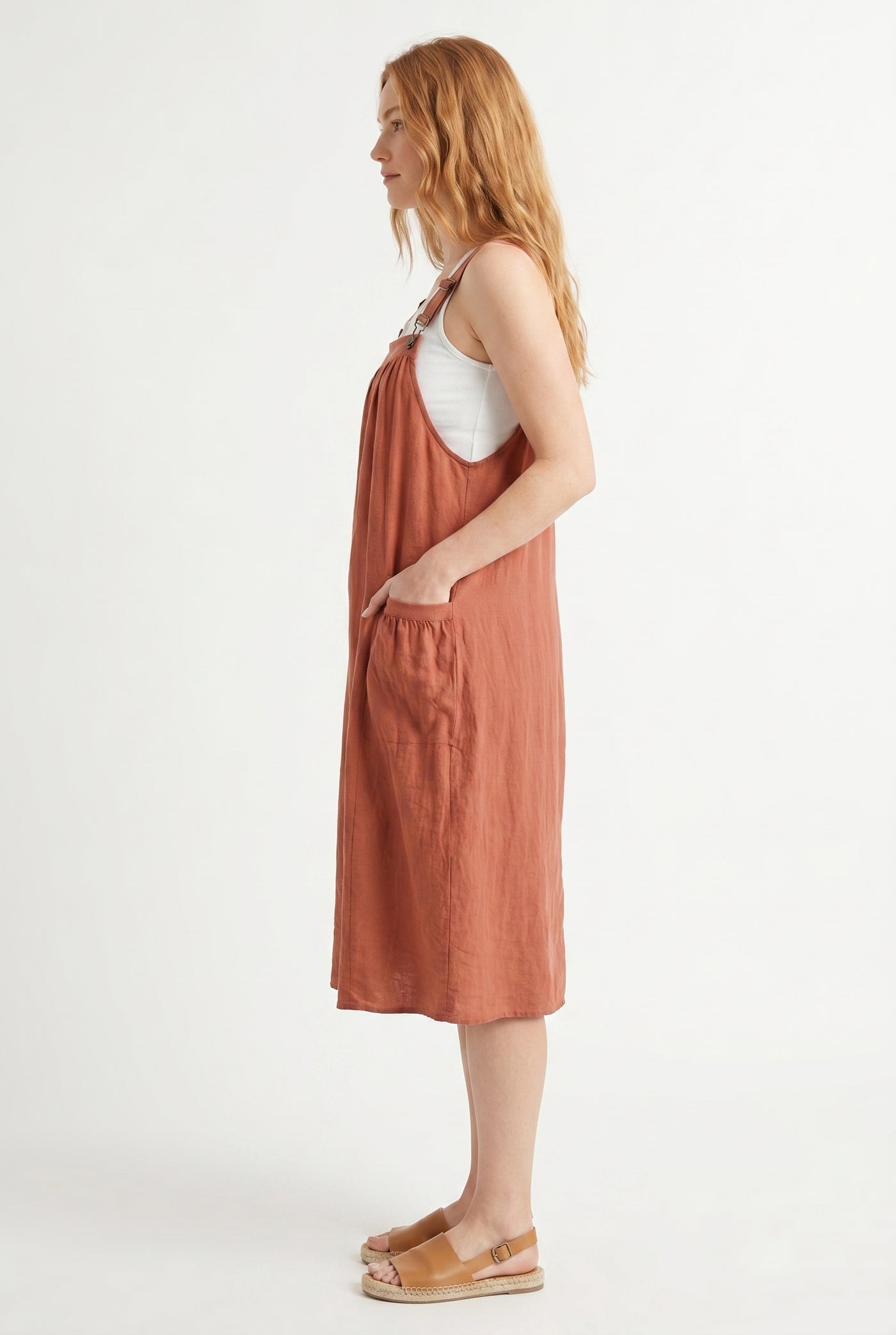Woman wearing a burnt sienna orange midi-length pinafore dress layered over a white long-sleeve top