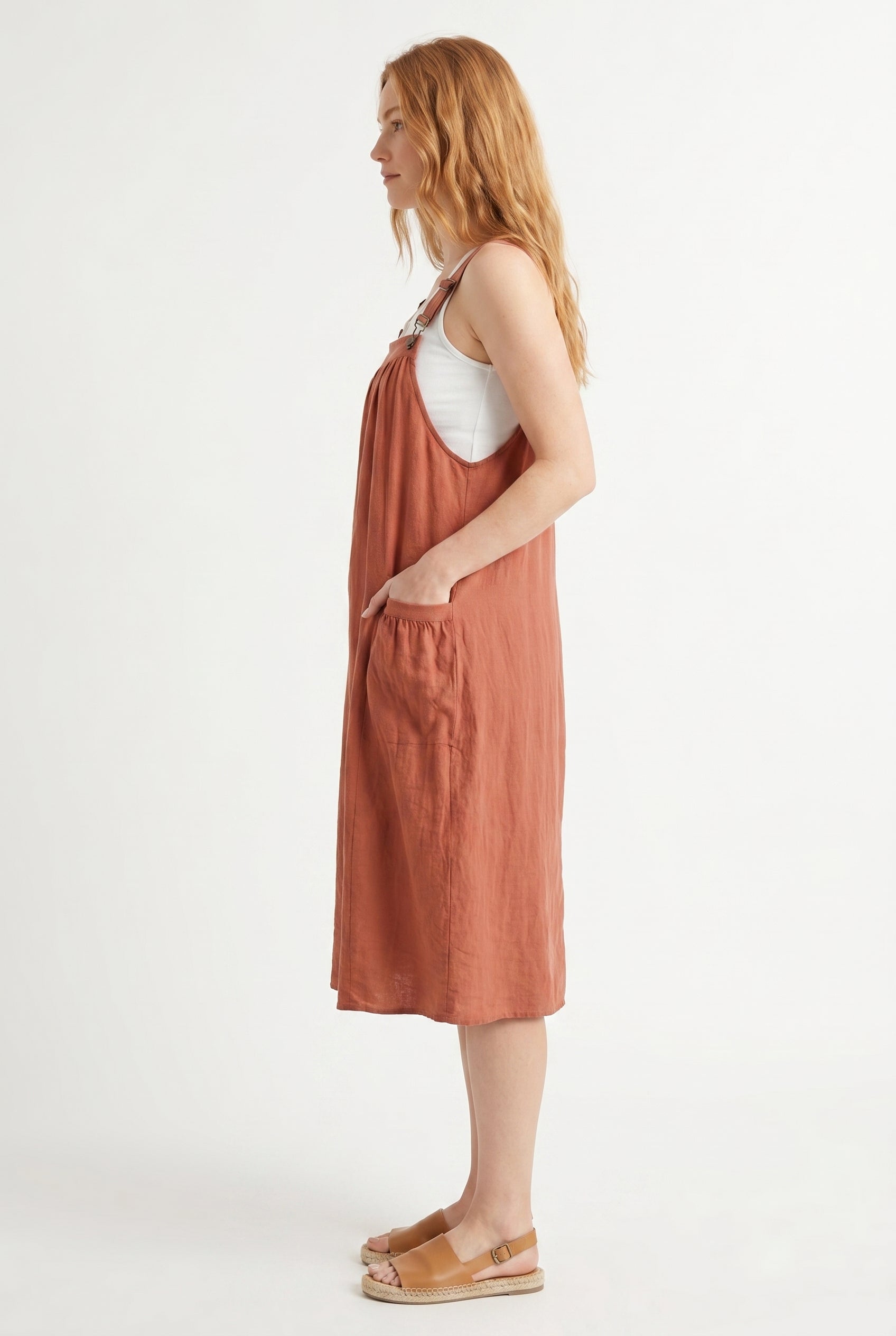 Woman wearing a burnt sienna orange midi-length pinafore dress layered over a white long-sleeve top
