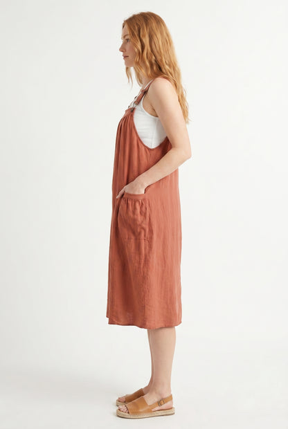 Woman wearing a burnt sienna orange midi-length pinafore dress layered over a white long-sleeve top