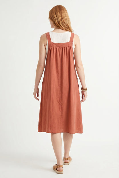 Woman wearing a burnt sienna orange midi-length pinafore dress layered over a white long-sleeve top