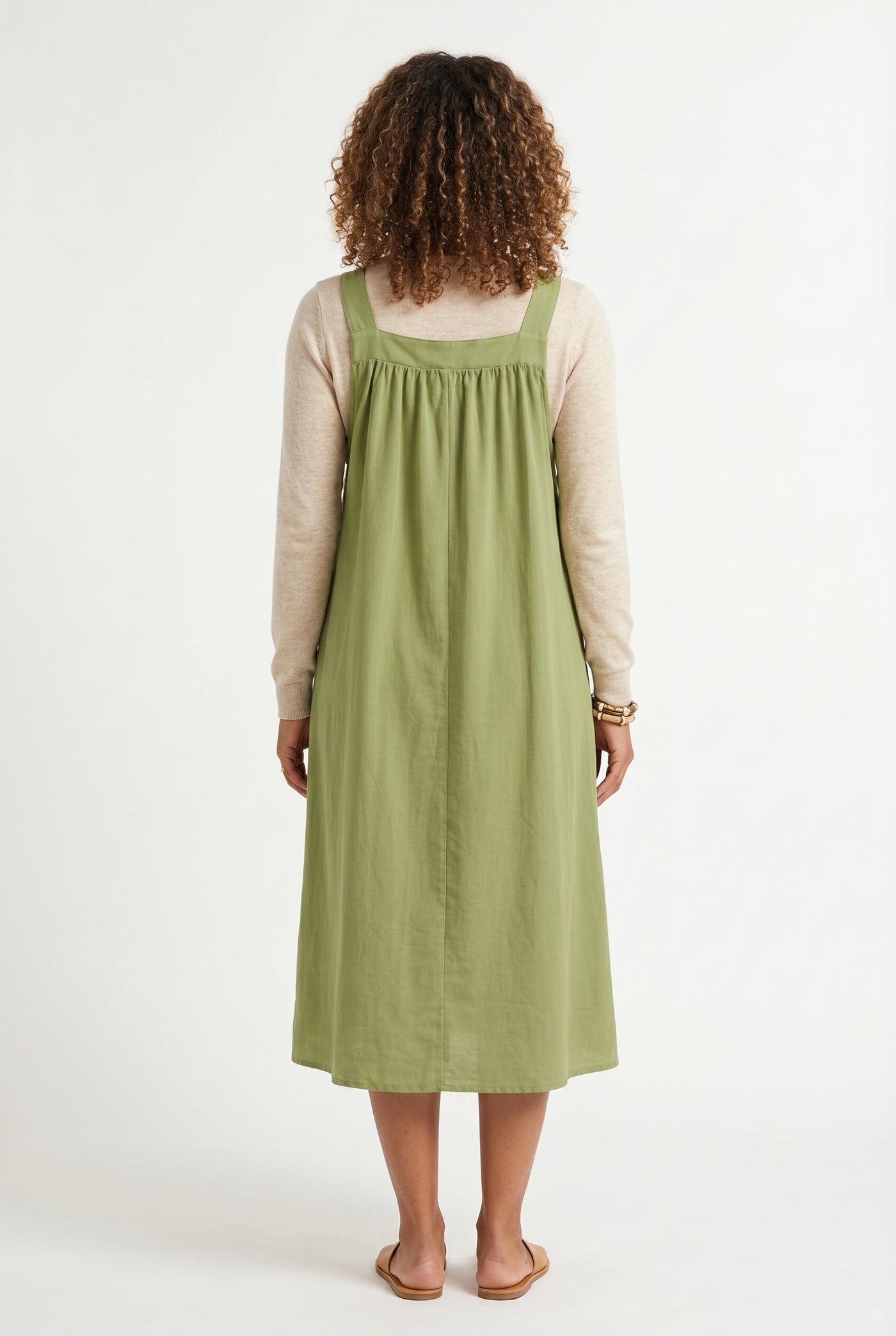 Woman wearing a matcha light green midi-length pinafore dress layered over a white long-sleeve top
