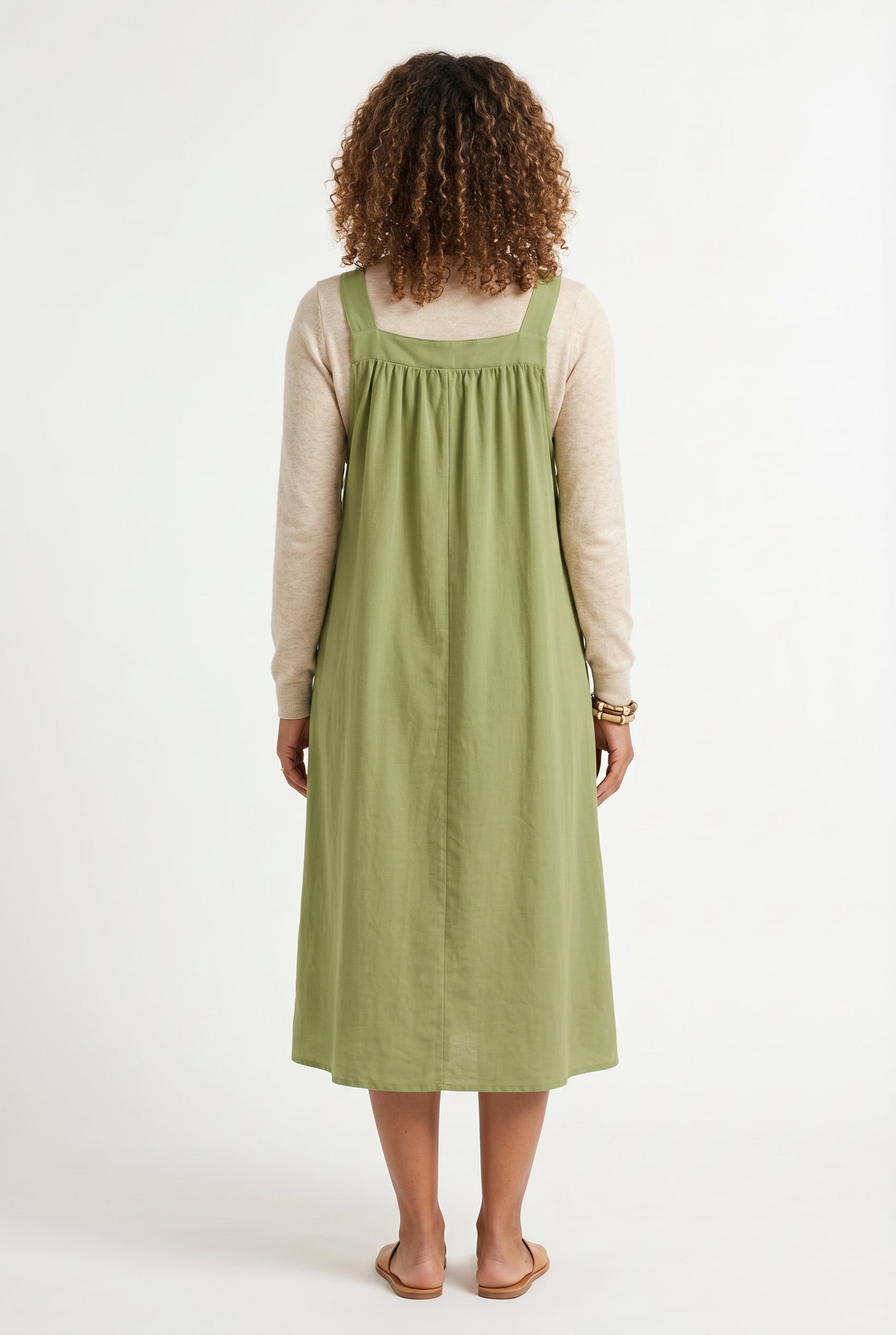 Woman wearing a matcha light green midi-length pinafore dress layered over a white long-sleeve top