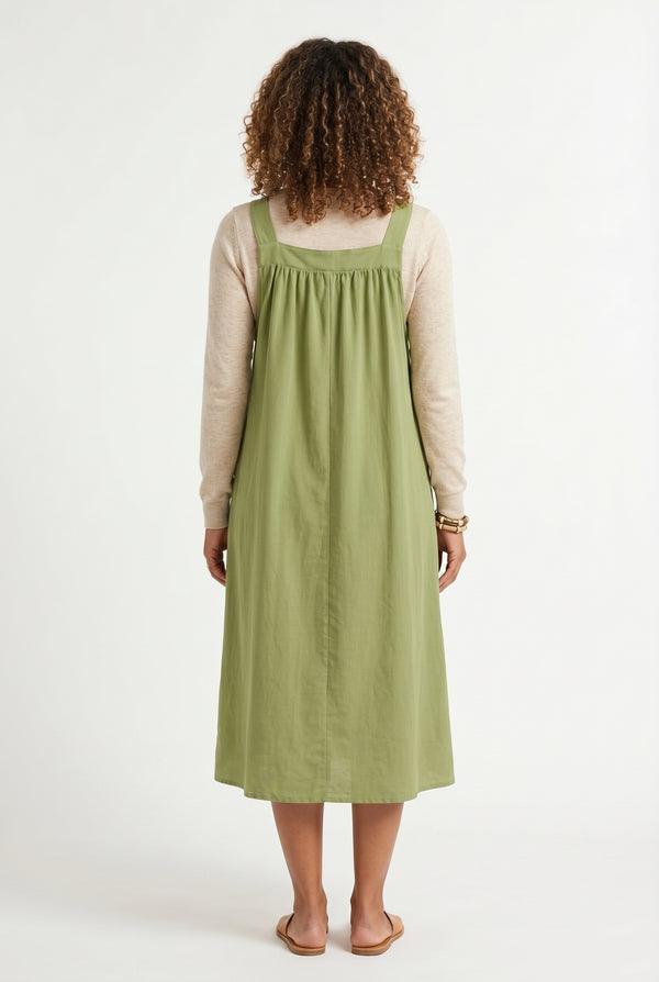Woman wearing a matcha light green midi-length pinafore dress layered over a white long-sleeve top