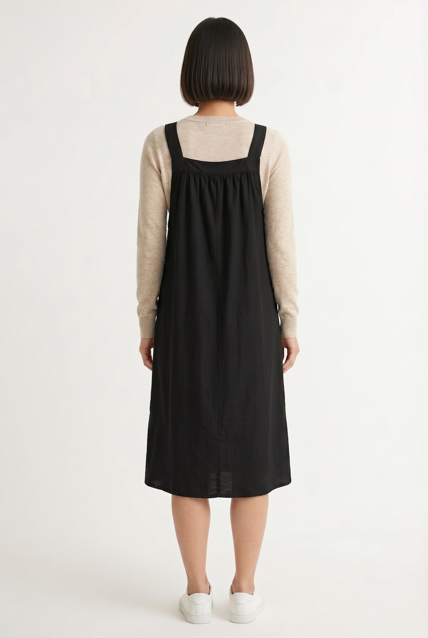 Woman wearing an onyx black midi-length pinafore dress layered over a white long-sleeve top