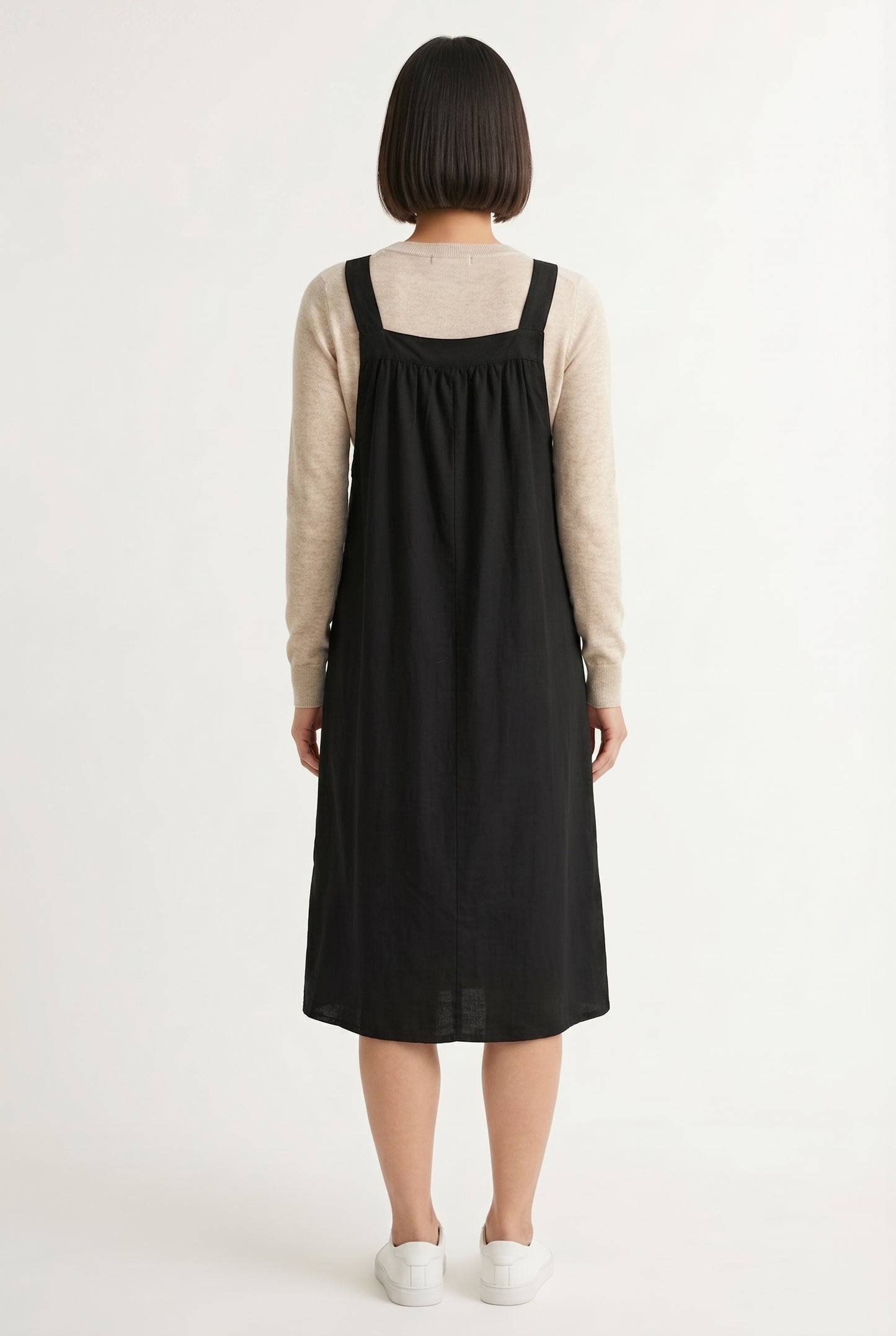 Woman wearing an onyx black midi-length pinafore dress layered over a white long-sleeve top