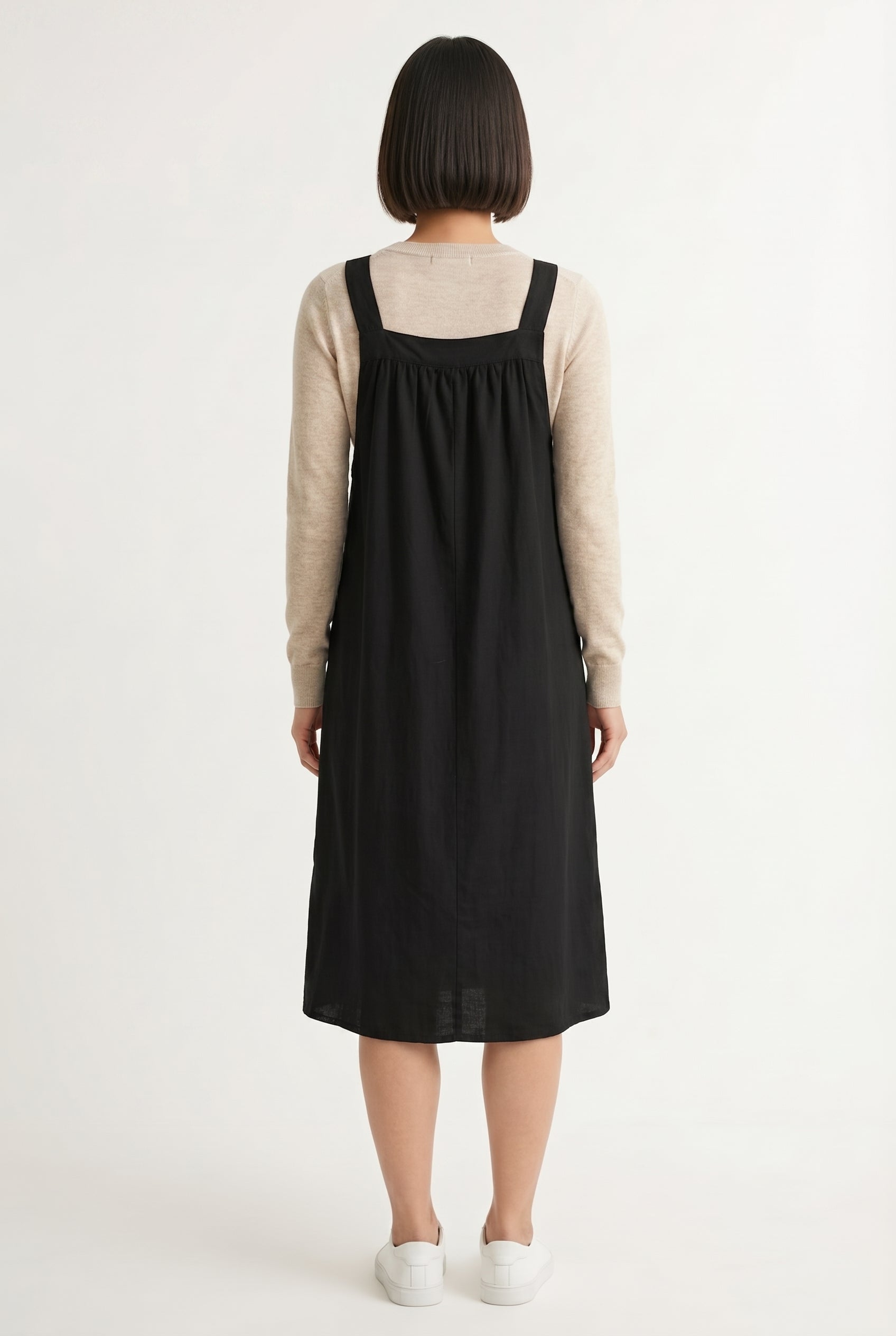 Woman wearing an onyx black midi-length pinafore dress layered over a white long-sleeve top