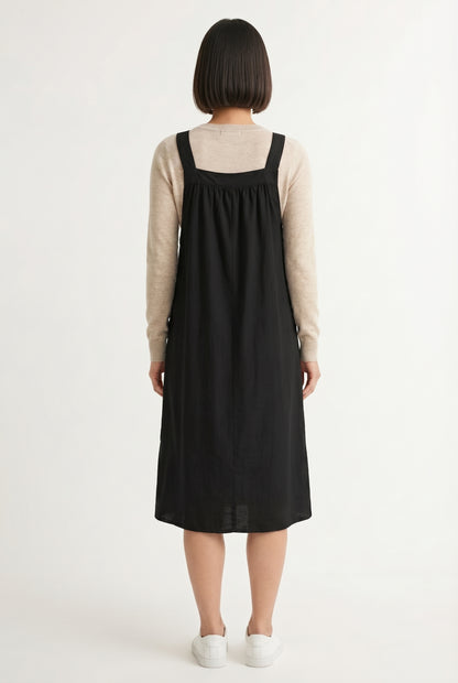 Woman wearing an onyx black midi-length pinafore dress layered over a white long-sleeve top