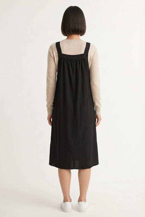 Woman wearing an onyx black midi-length pinafore dress layered over a white long-sleeve top