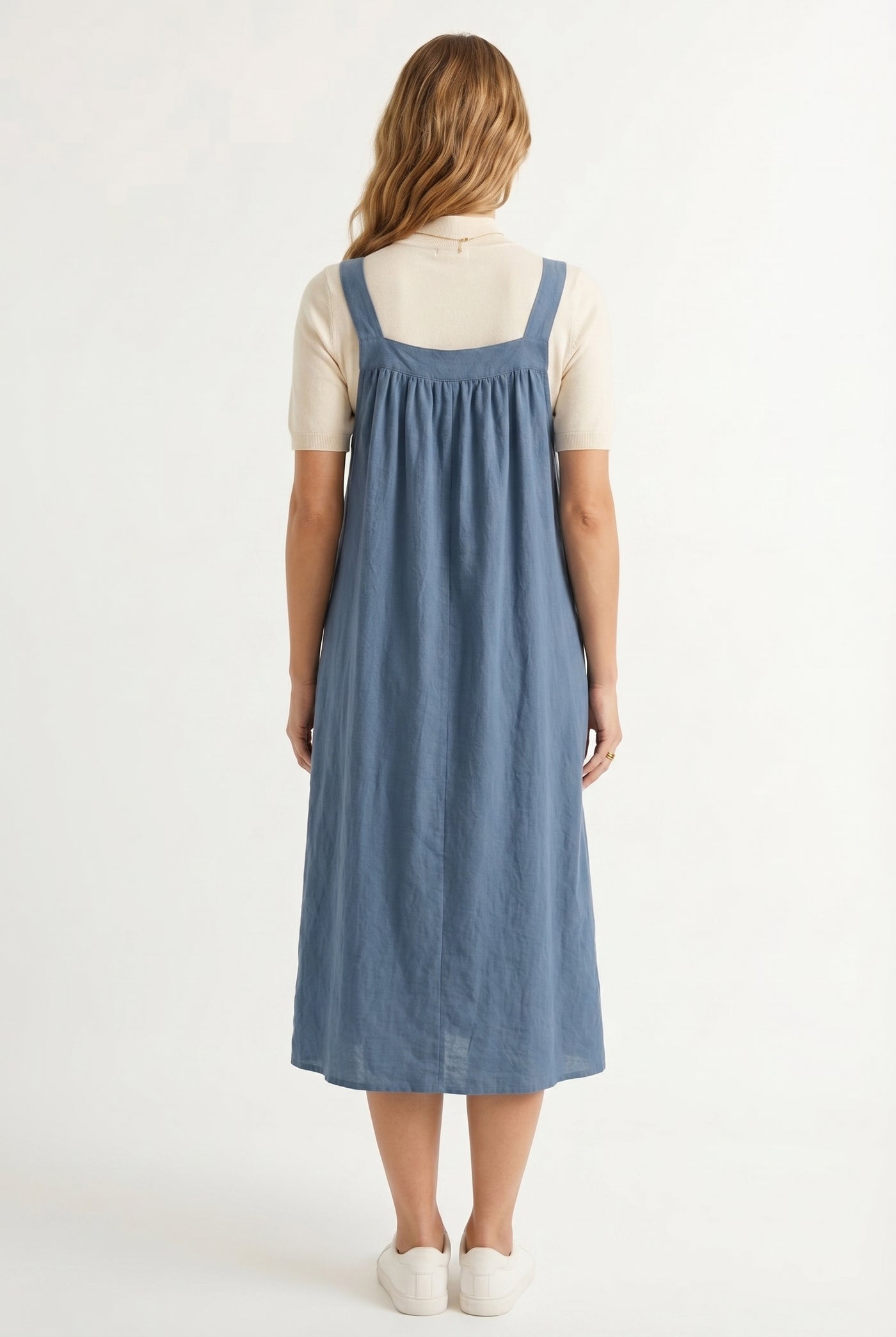 Woman wearing a storm blue midi-length pinafore dress layered over a white long-sleeve top