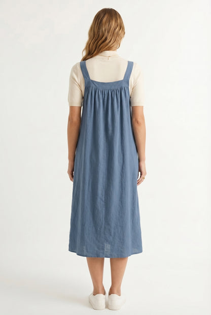 Woman wearing a storm blue midi-length pinafore dress layered over a white long-sleeve top