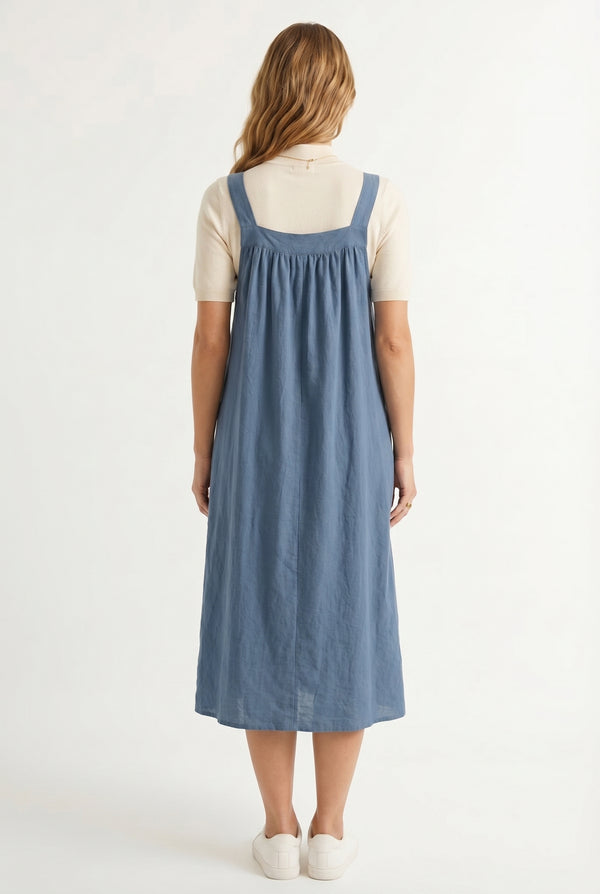 Woman wearing a storm blue midi-length pinafore dress layered over a white long-sleeve top