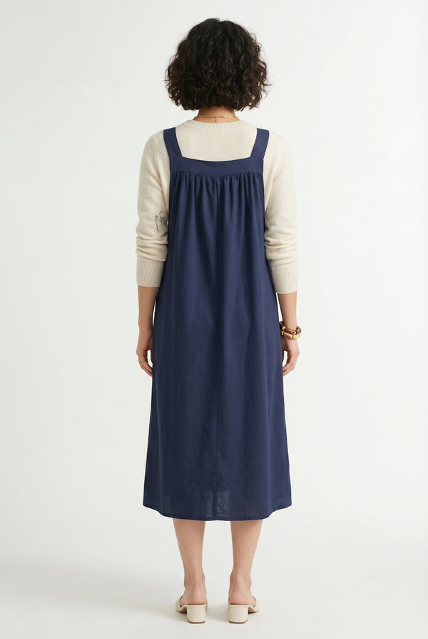 Woman wearing a total eclipse dark navy blue midi-length pinafore dress layered over a white long-sleeve top