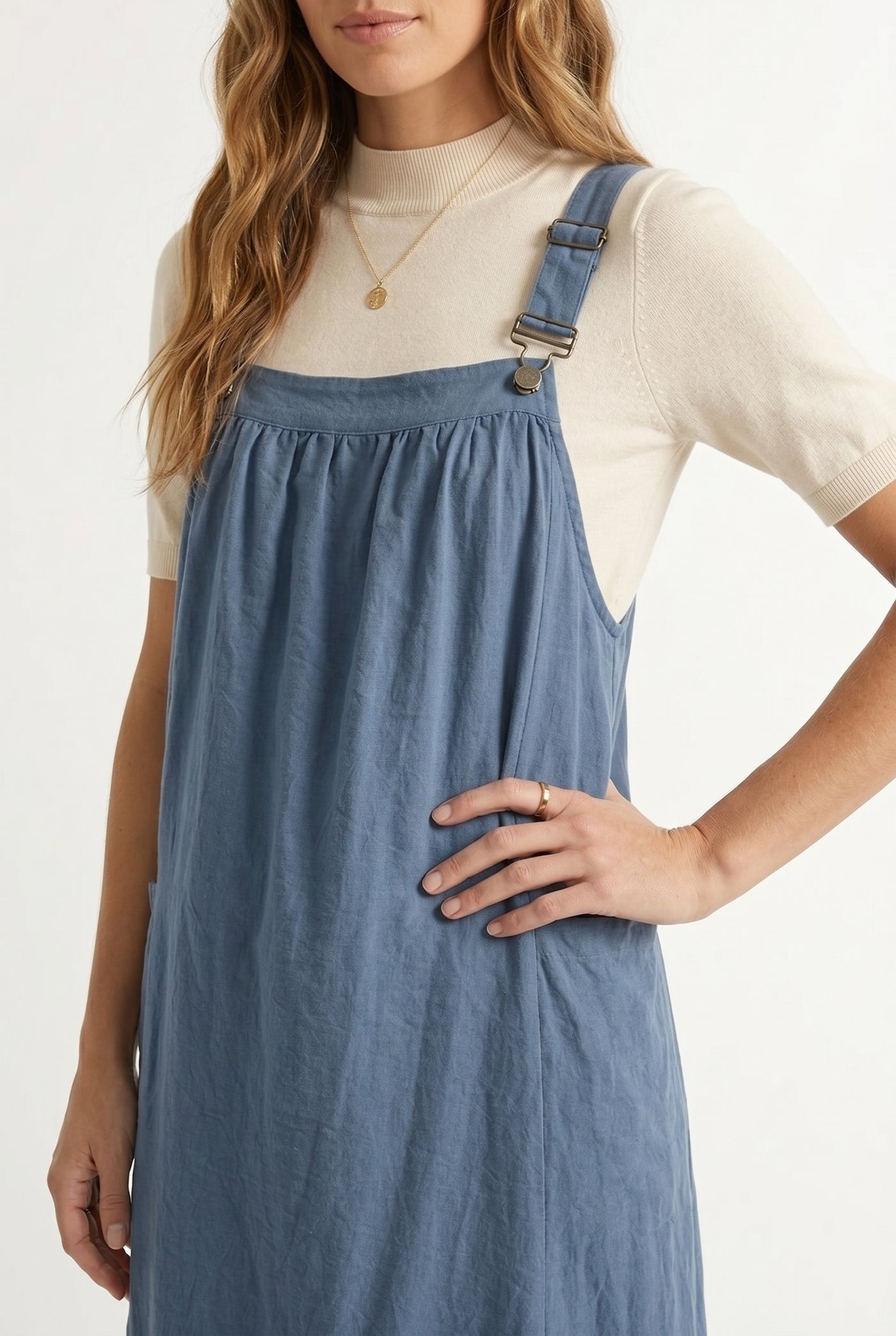 Woman wearing a storm blue midi-length pinafore dress layered over a white long-sleeve top