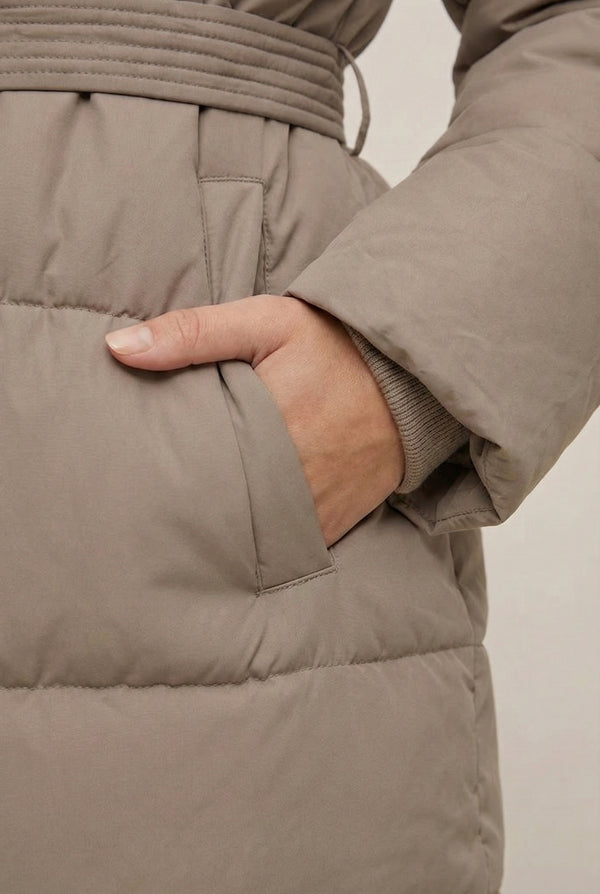 Woman modeling the belted Aspen puffer jacket in Fawn by LOHNR, featuring a detachable faux fur hood.