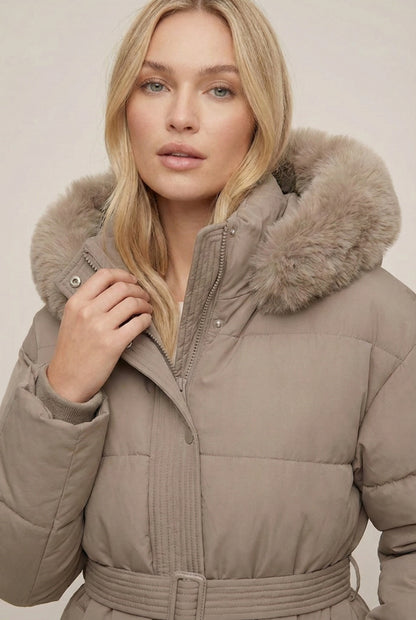Woman modeling the belted Aspen puffer jacket in Fawn by LOHNR, featuring a detachable faux fur hood.