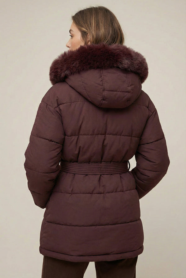 Woman modeling the belted Aspen puffer jacket in Merlot by LOHNR, featuring a detachable faux fur hood.