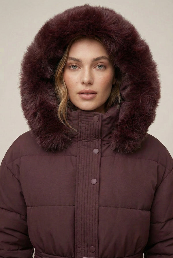Woman modeling the belted Aspen puffer jacket in Merlot by LOHNR, featuring a detachable faux fur hood.