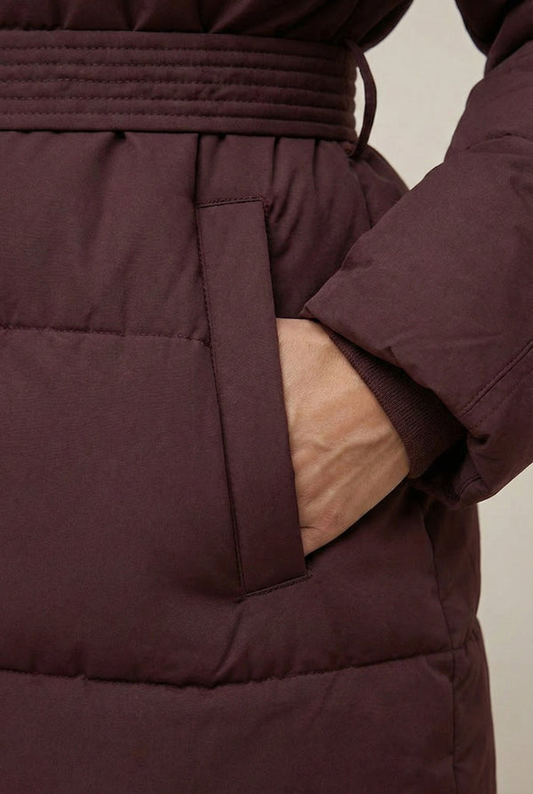 Woman modeling the belted Aspen puffer jacket in Merlot by LOHNR, featuring a detachable faux fur hood.