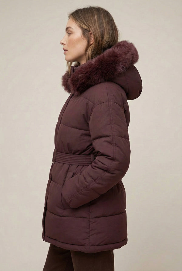 Woman modeling the belted Aspen puffer jacket in Merlot by LOHNR, featuring a detachable faux fur hood.