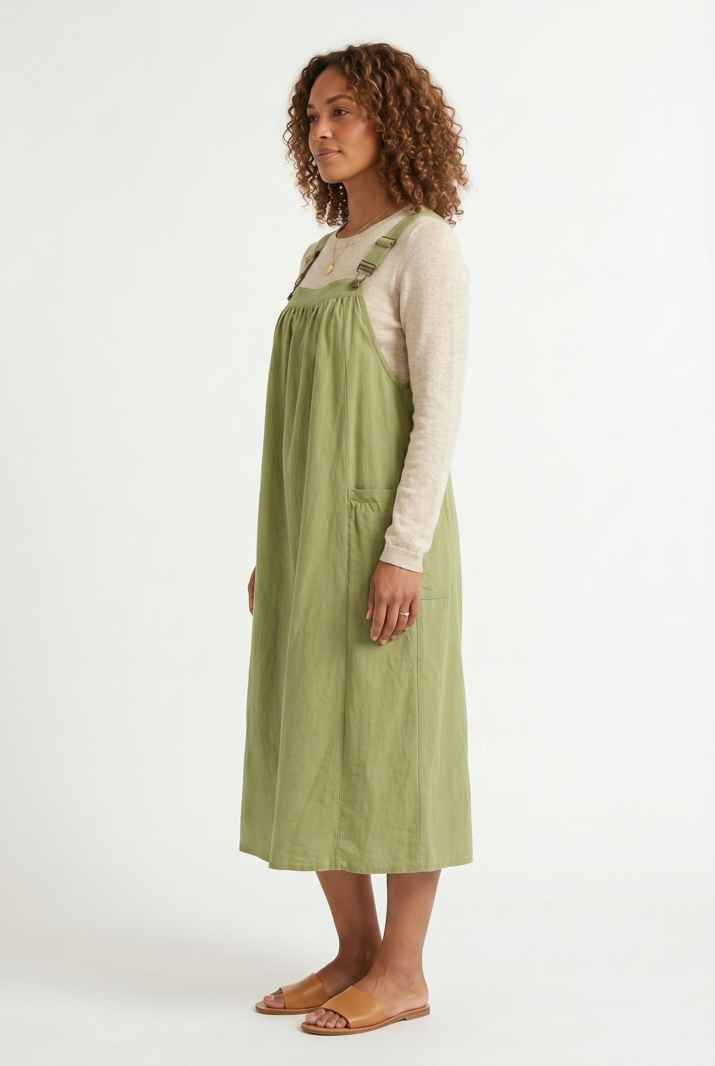 Woman wearing a matcha light green midi-length pinafore dress layered over a white long-sleeve top