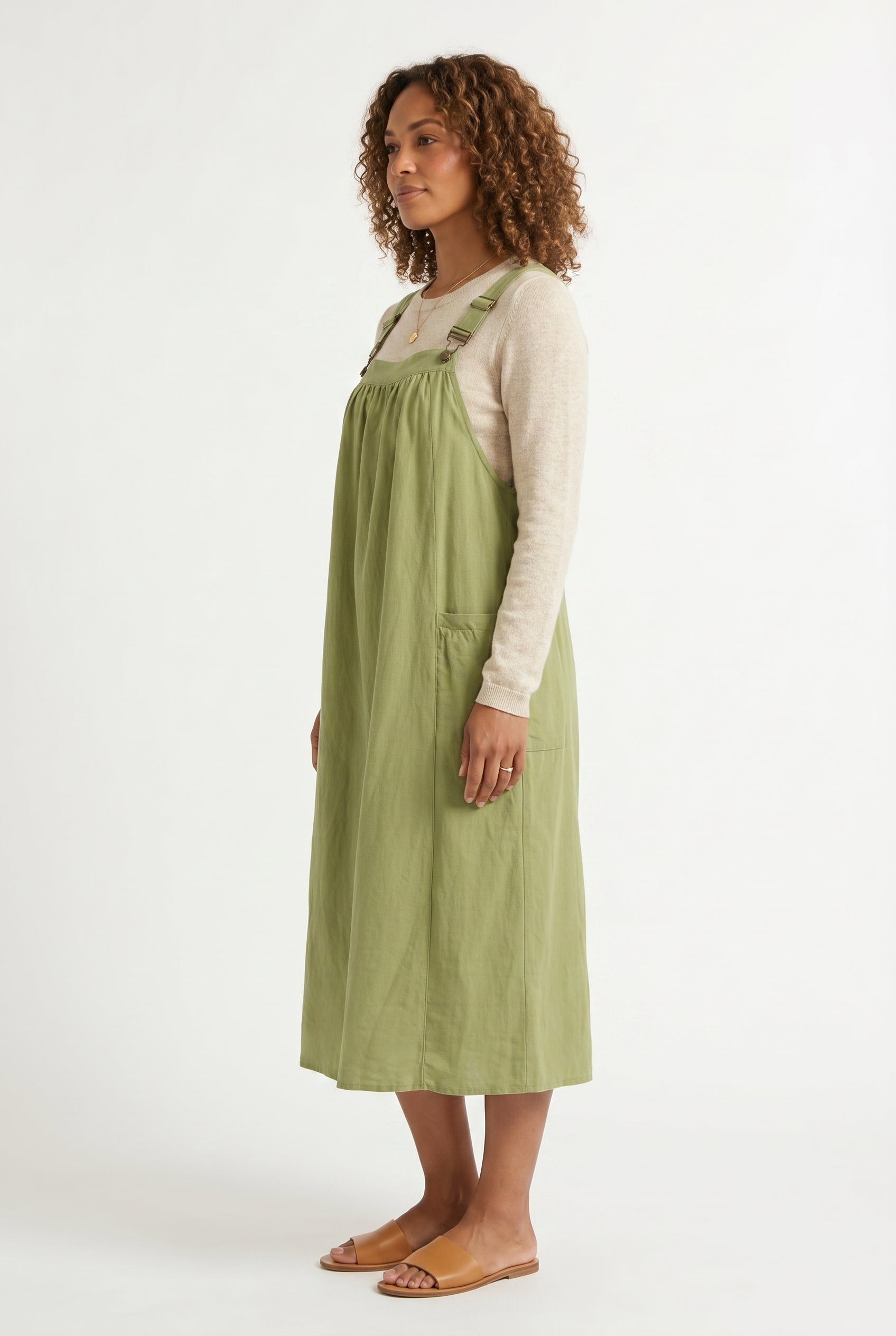 Woman wearing a matcha light green midi-length pinafore dress layered over a white long-sleeve top