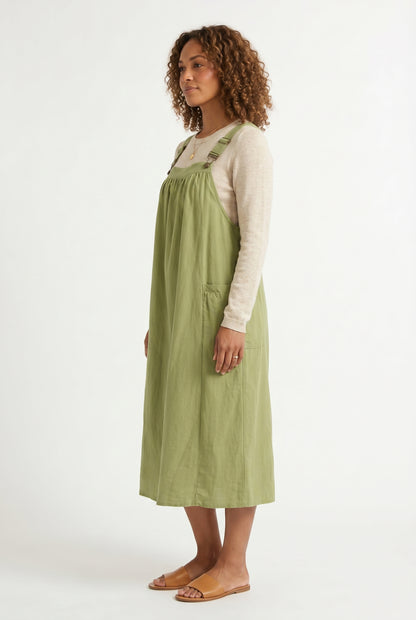 Woman wearing a matcha light green midi-length pinafore dress layered over a white long-sleeve top