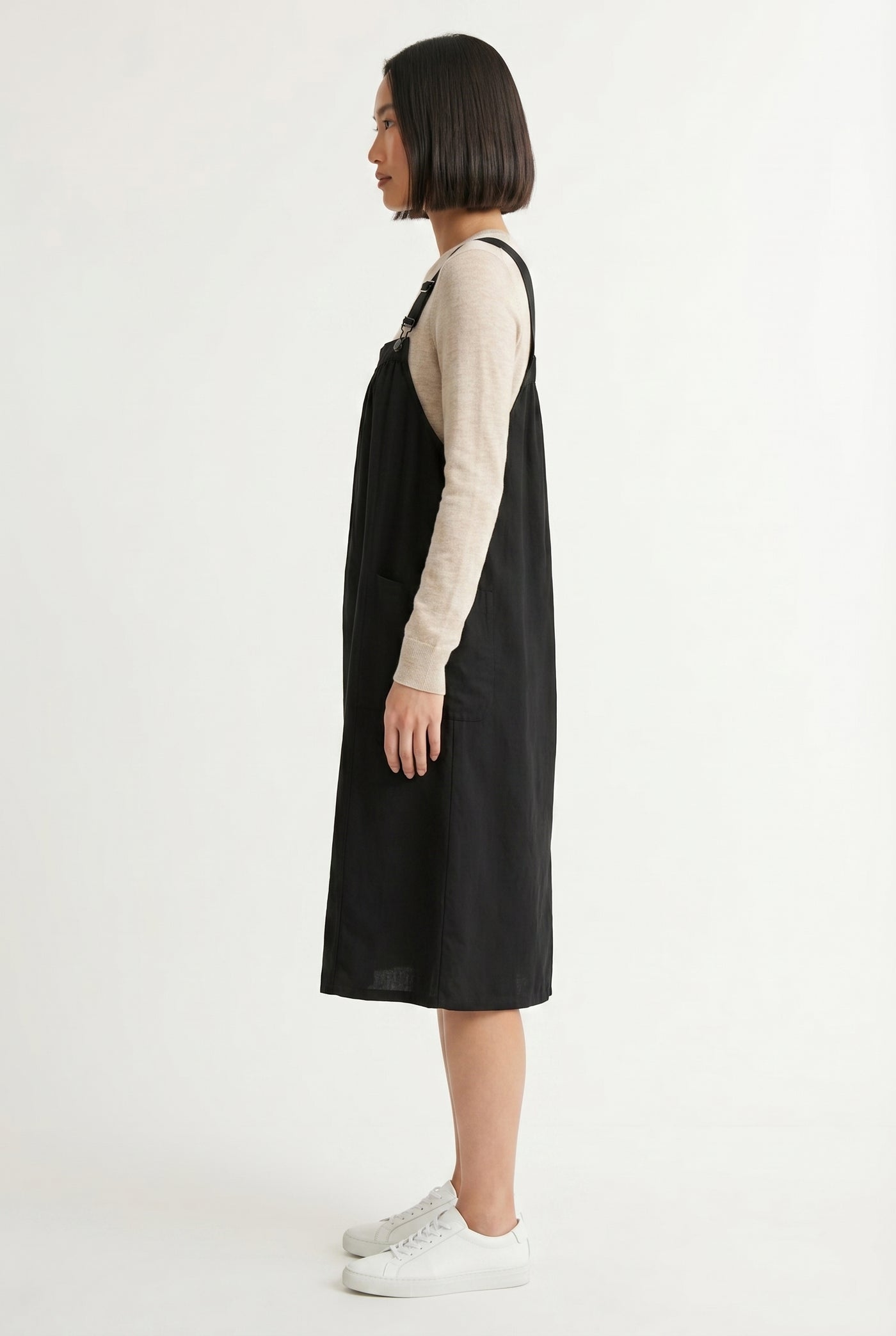 Woman wearing an onyx black midi-length pinafore dress layered over a white long-sleeve top