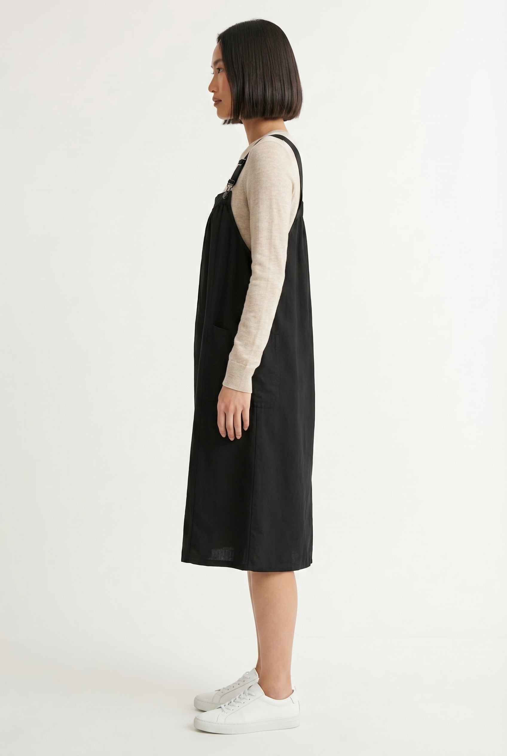 Woman wearing an onyx black midi-length pinafore dress layered over a white long-sleeve top