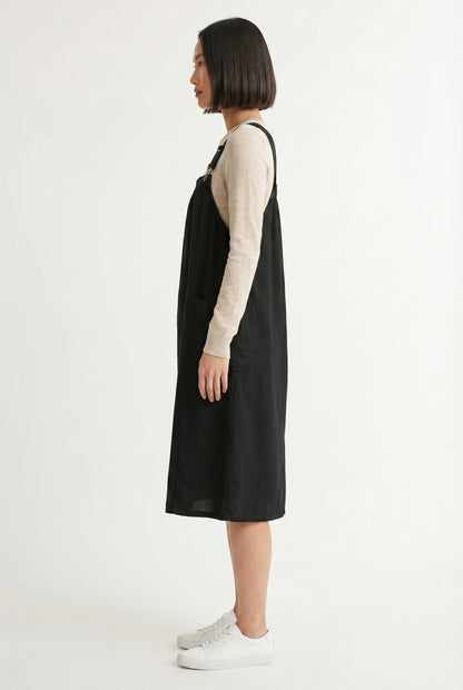 Woman wearing an onyx black midi-length pinafore dress layered over a white long-sleeve top