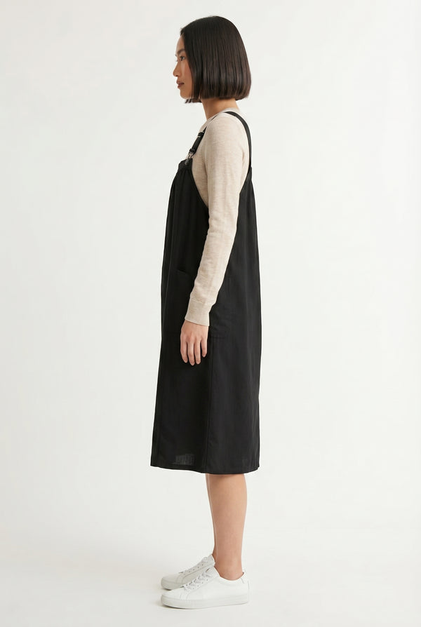 Woman wearing an onyx black midi-length pinafore dress layered over a white long-sleeve top