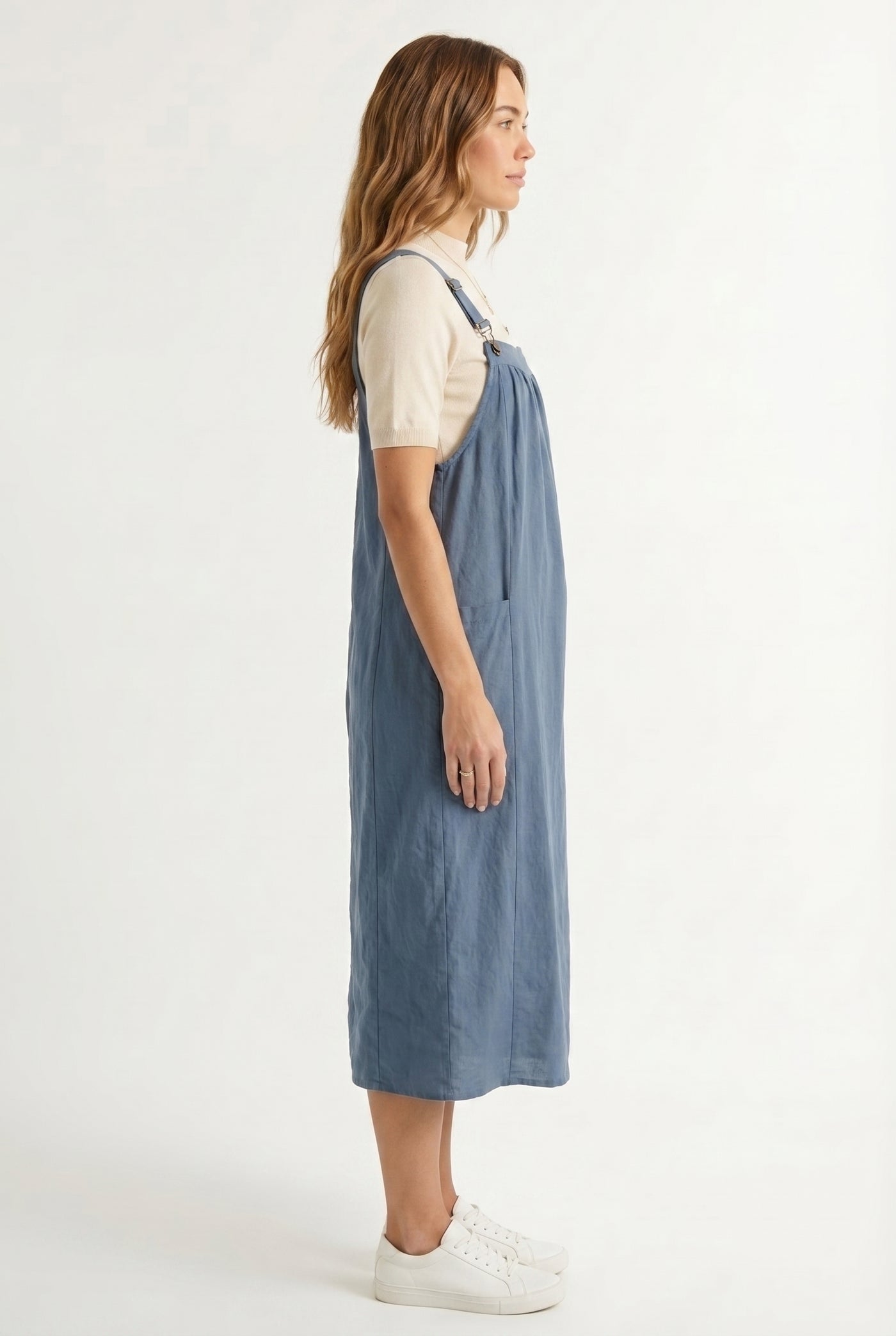 Woman wearing a storm blue midi-length pinafore dress layered over a white long-sleeve top