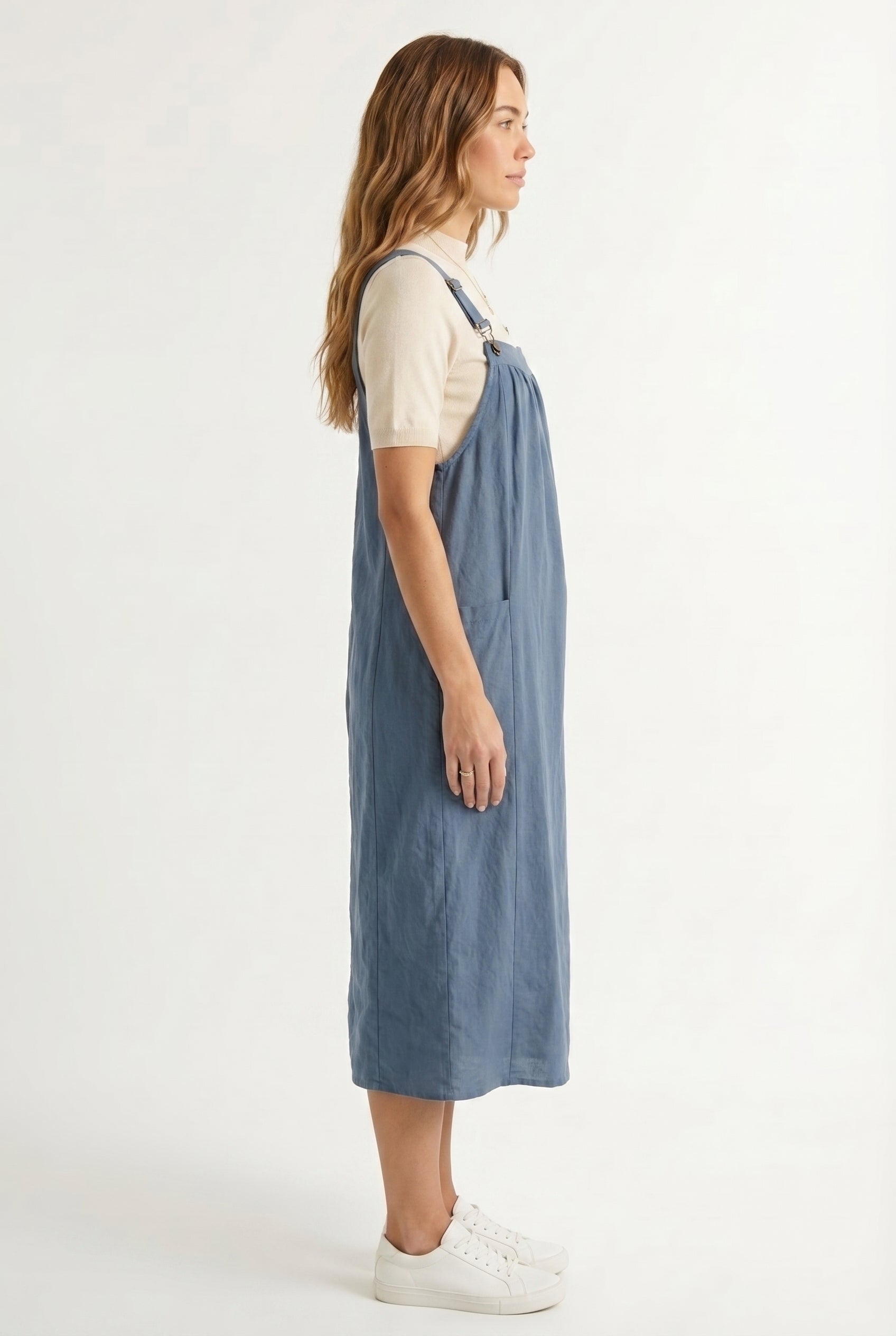 Woman wearing a storm blue midi-length pinafore dress layered over a white long-sleeve top